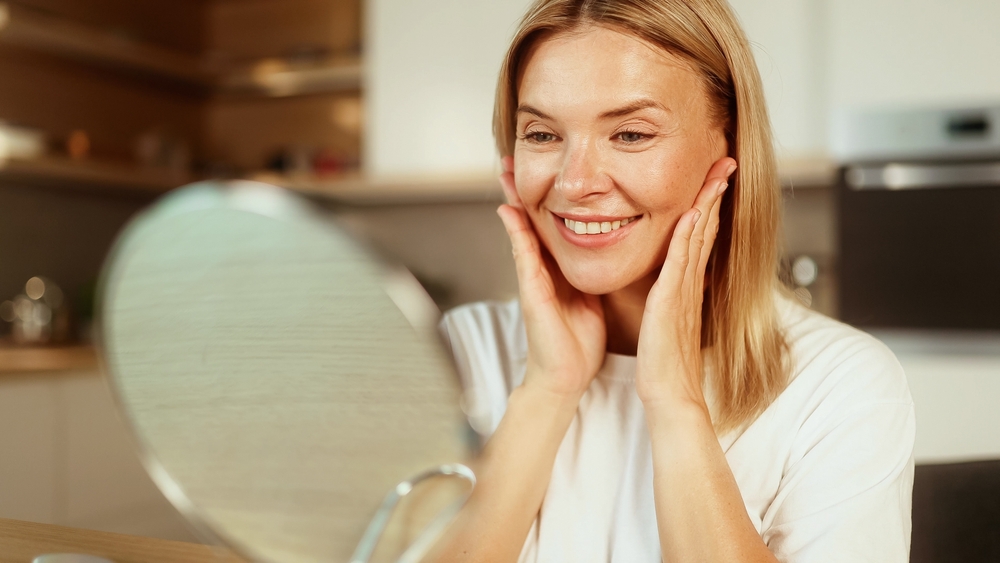 Smiling woman examining radiant skin in mirror after collagen therapy